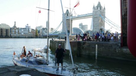 Segelyacht mit drei Personen an Bord liegt an einem Anleger auf der Themse in London, unmittelbar vor der Tower Bridge. Zahlreiche Zuschauer beobachten das Boot vom Kai aus.